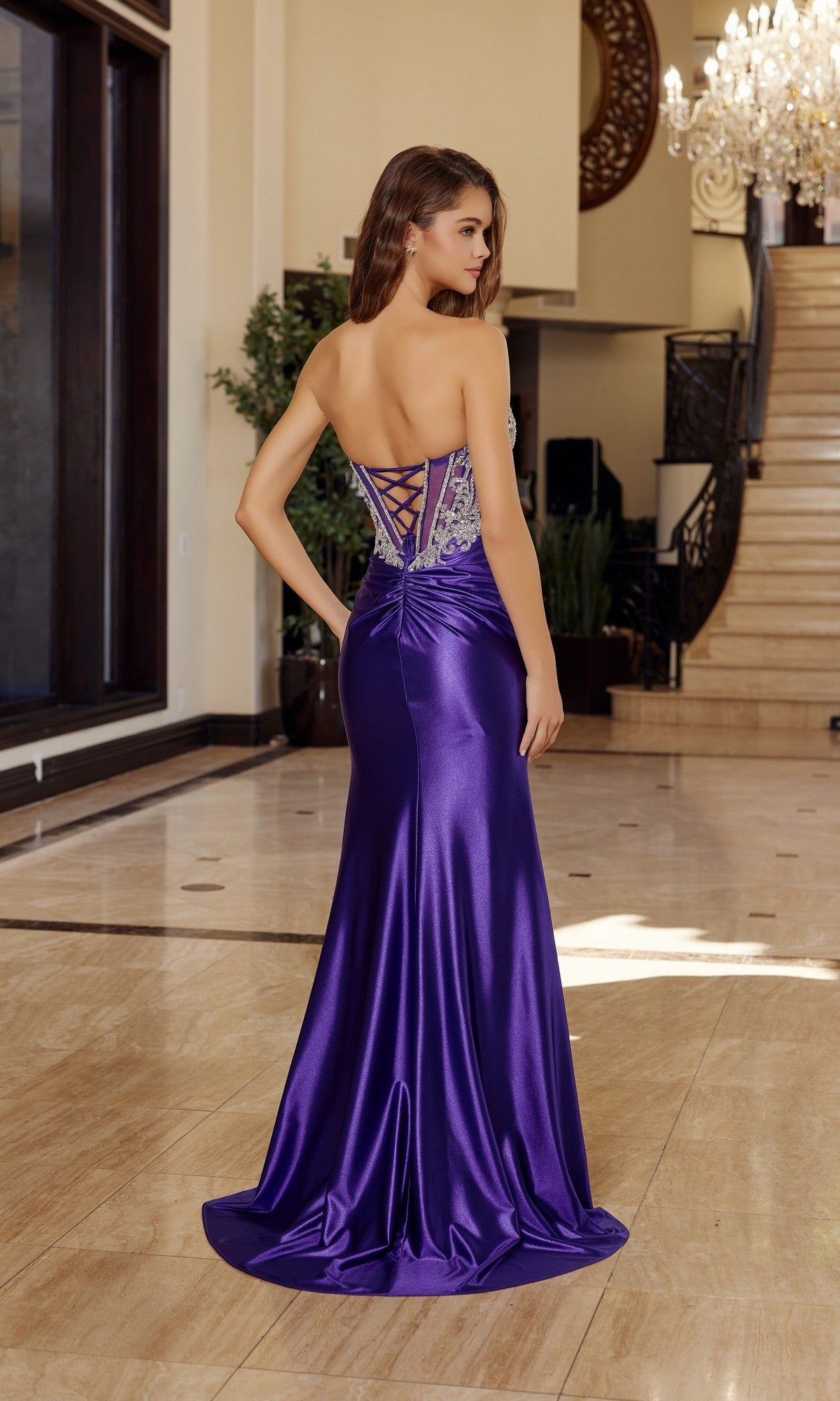 A woman in the Nox Anabel T1854, a strapless satin long prom dress with a lace-up back, stands indoors near a staircase as her floor-length purple gown catches the light.